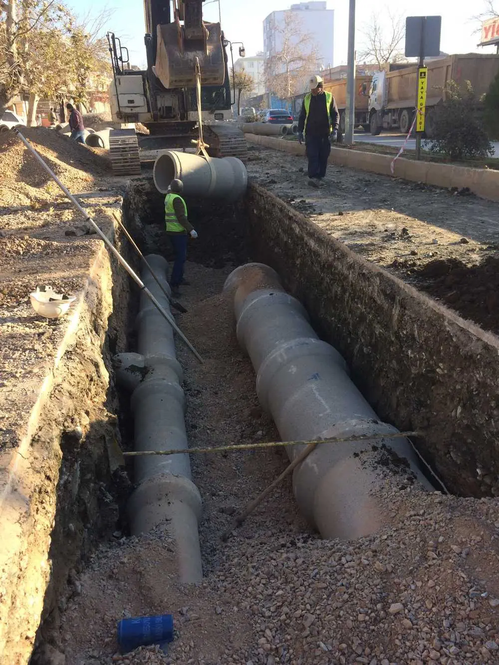 Kentsel alanda hendek kazısı içinde iki sıra beton drenaj borusu döşeme işlemi; ekskavatör ile boru indirme, reflektif yelek giyen işçiler, yol kenarı şantiye