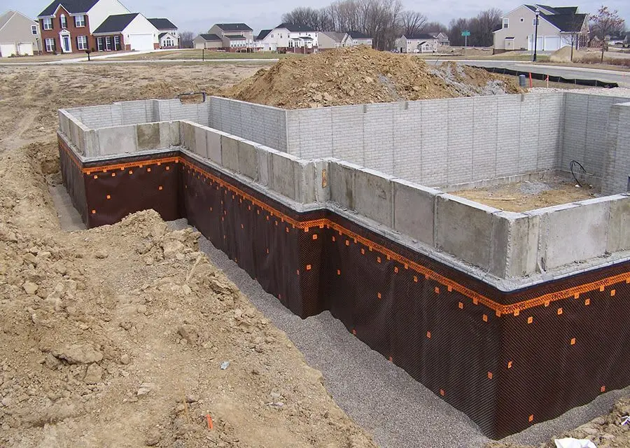 İnşaat halinde bir bodrum katı dışarıdan görünüm: betonarme perde duvarların dış yüzeyine kahverengi/turuncu noplu HDPE drenaj levhaları monte edilmiş; temel tabanı seviyesinde agrega dolgu ve zemin hazırlığı yapılmaktadır