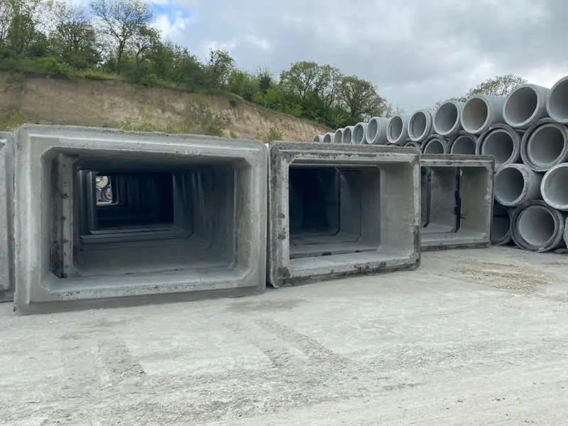 Saha stok alanında yan yana dizilmiş prefabrik betonarme kutu menfez elemanları (dikdörtgen kesitli, açık uçlu) ve arka planda istiflenen dairesel beton boru menfez elemanları