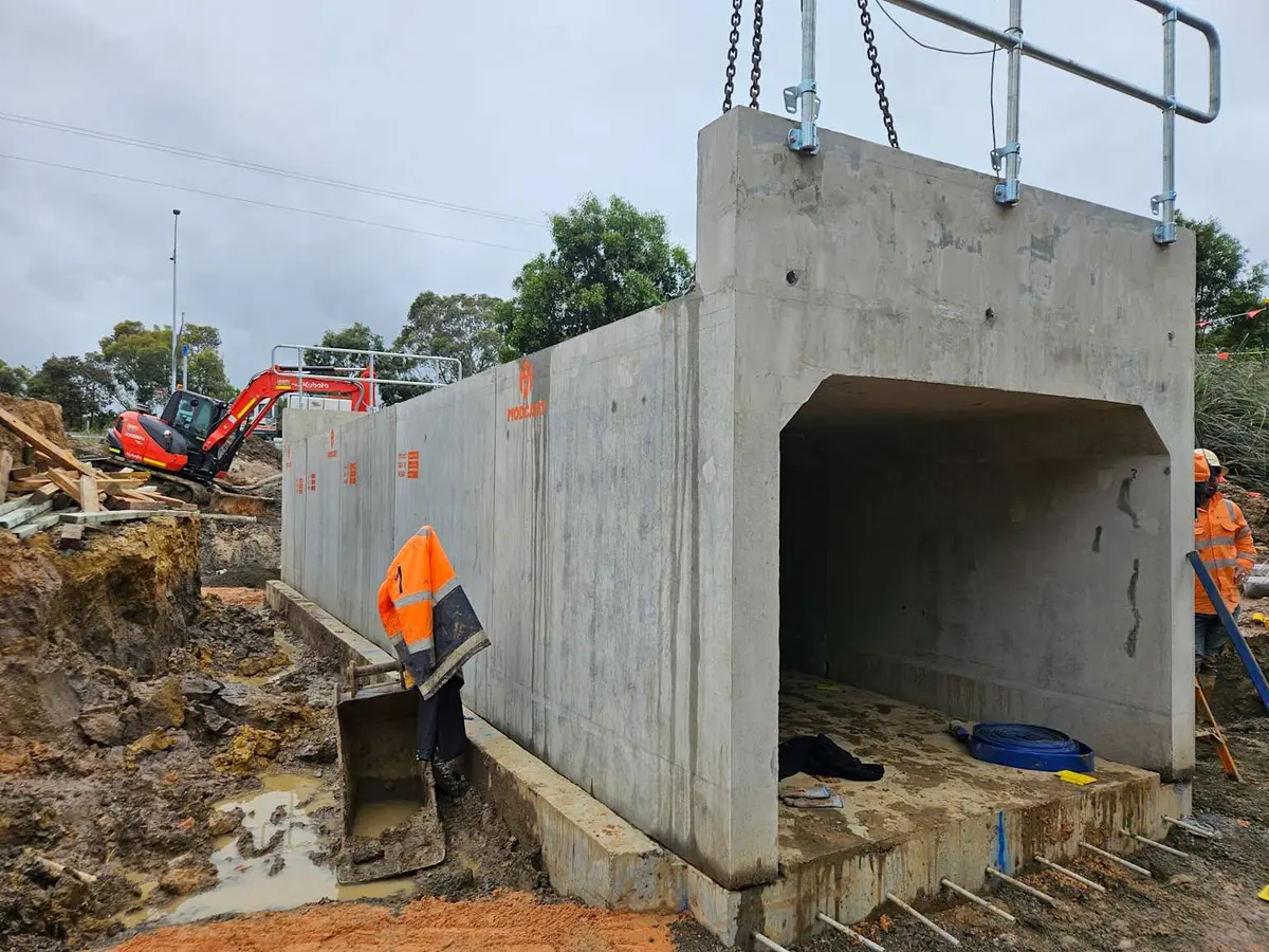 Büyük bir prefabrik betonarme kutu menfez elemanının vinçle trençe indirildiği saha montajı; işçiler kanat duvarları ve montaj ankrajlarını hazırlamaktadır