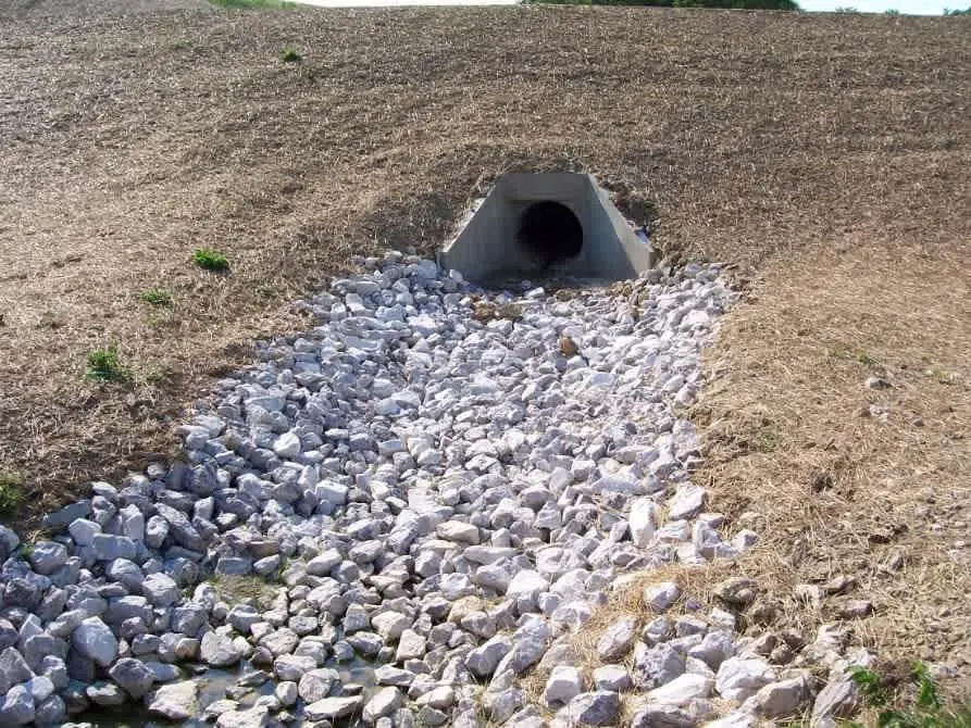 Tarım arazisi içindeki yol dolgusu altından geçen boru menfez çıkışında mansap erozyon koruması için döşenmiş beyaz kırma taş rip-rap apron; menfez çıkışından mansap kanalına doğru yayılan kama biçimli taş koruma görülmektedir