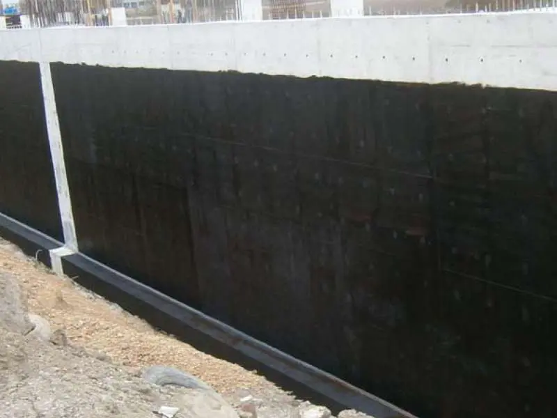 İnşaat sahası bodrum perde duvarına bitümlü su yalıtım membranı uygulanmış uzun kesit görünümü — siyah bitüm kaplı betonarme perde, üstte donatı çerçevesi, altta hazırlık çakılı