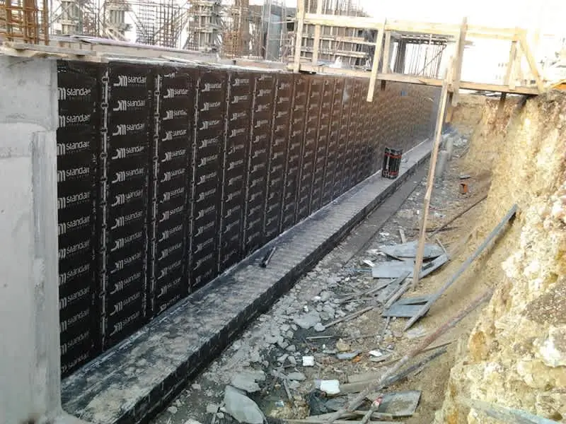 Standar markalı HDPE profillenmiş drenaj levhalarının bodrum perde duvarı boyunca montajı — desenli siyah levhalar art arda tutturulmuş, arkaplanda kaya-toprak yüzey