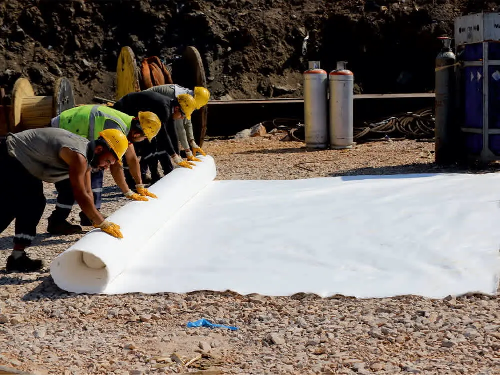 Birkaç işçi çakıl zemin üzerinde beyaz nonwoven geotekstil rulosunu birlikte açıyor — sarı baret ve güvenlik yelekli çalışanlar