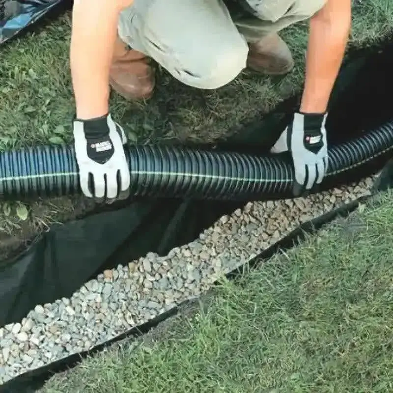 Eldivenli işçi geotekstil sarılı siyah HDPE delikli boruyu hendek içine yerleştiriyor — altında beyaz çakıl, açık siyah geotekstil şerit