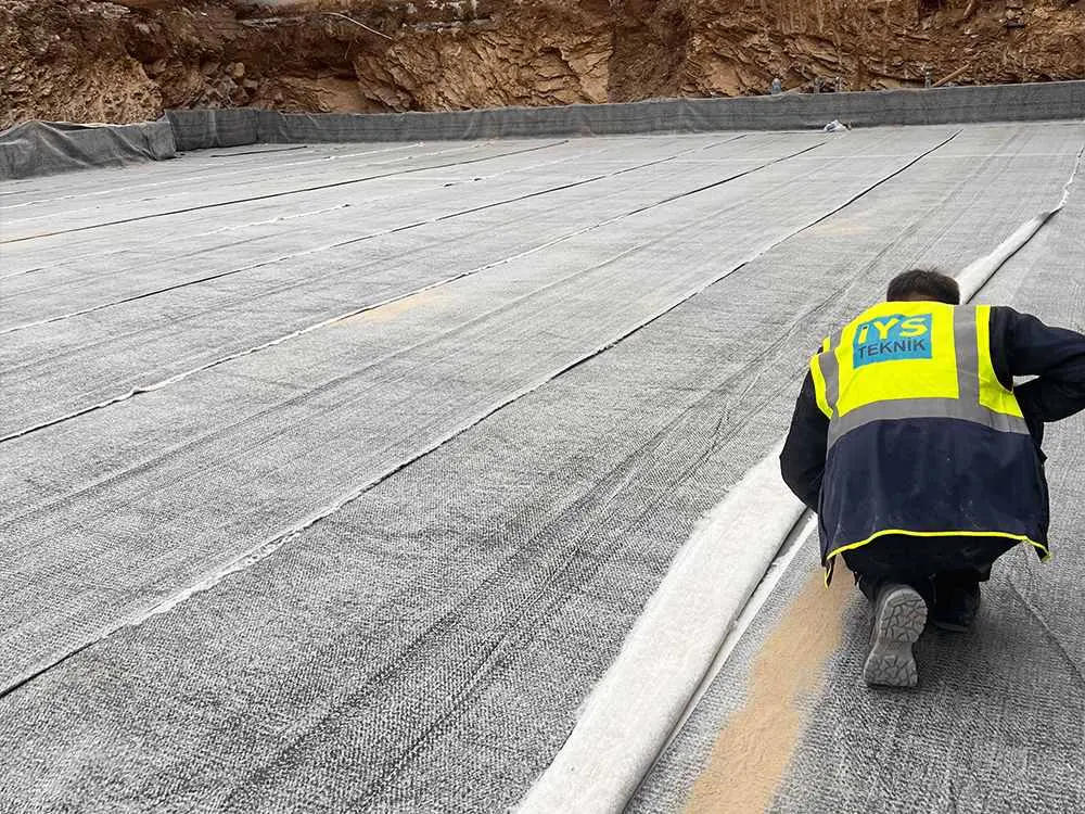 LYS TEKNİK logolu sarı yelek giyen işçi gri nonwoven geotekstil serimini denetliyor — açık yüzey üzerinde paralel şeritler halinde serilmiş geniş geotekstil