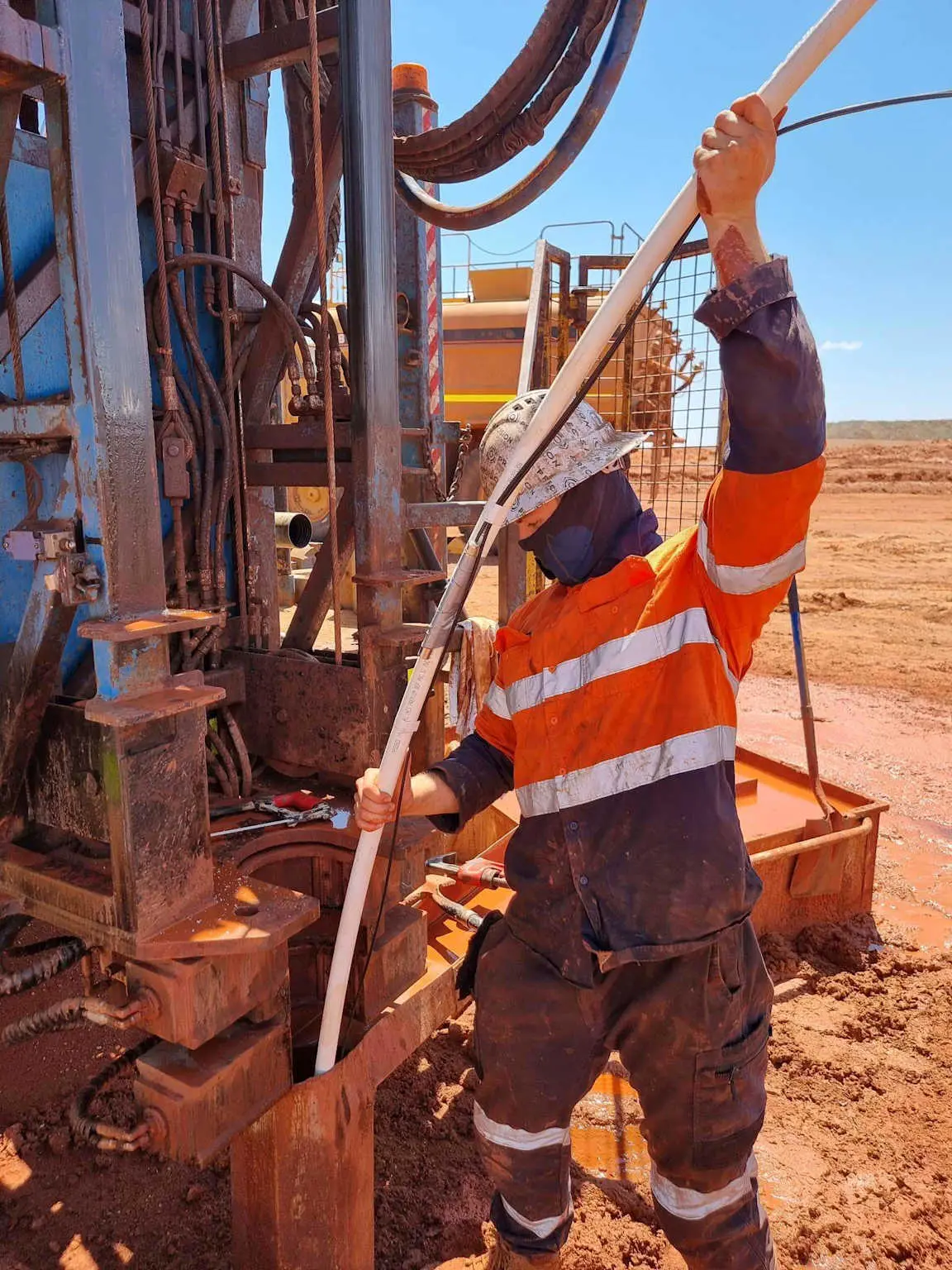 Sondaj kulesi yanında turuncu-sarı güvenlik yelekli işçi sondaj borulaması yapıyor — kırmızımsı toprak sahası, arka planda sondaj makinesi ekipmanları