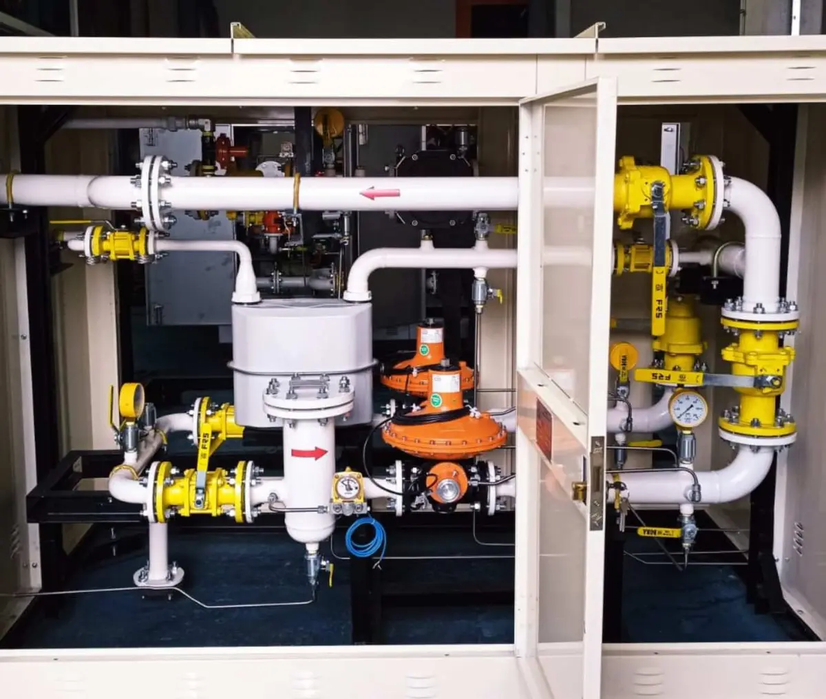 Doğal gaz basınç düşürme ve ölçüm istasyonu (RDIM) yakın görünüm — sarı el vanaları, turuncu düzenleyici (regülatör), gri filtre, manometre ve bağlantı boruları