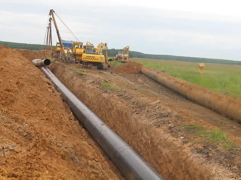 Büyük çaplı çelik doğal gaz iletim boru hattı açık arazide geniş hendek döşemesi — ön planda dış kaplı siyah boru, arka planda Komatsu vinç ve ekskavatör iş makineleri