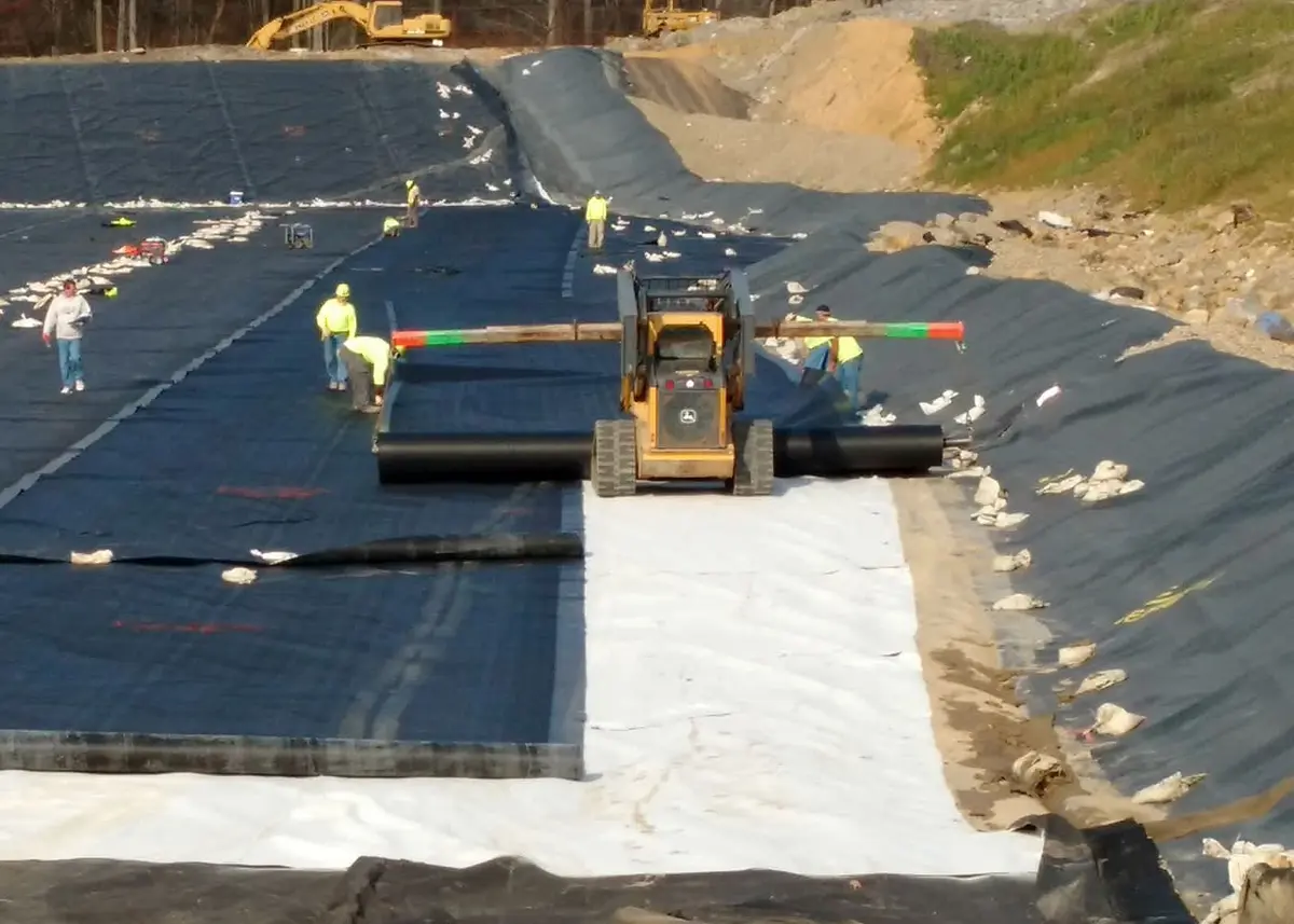 Düzenli depolama sahası taban liner montajı: siyah HDPE geomembran üzerine beyaz geotekstil tabakası açılıyor; sarı yansıtıcı yelekli işçiler ve John Deere ekskavatör yardımıyla geniş şev ve taban yüzeyi kaplanıyor