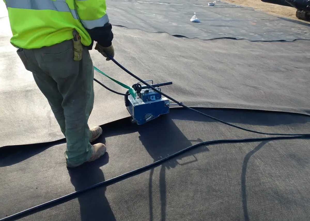 Saha kaynak uygulaması: sarı yansıtıcı yelekli kaynak teknisyeni çift sıcak takoz kaynak aleti ile siyah HDPE geomembran üzerinde dikiş hattı boyunca ilerliyor; günlük kalite kontrol testi öncesi işlem