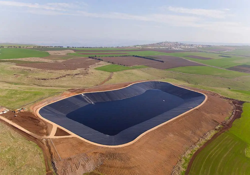 Tarım arazisi ortasında oval şekilli büyük sulama/sulama rezervuarı havadan drone görünümü: siyah HDPE geomembran ile tamamen kaplanmış su deposu, etrafında kırmızı toprak, yeşil ekilmiş tarlalar ve uzakta yerleşim alanı