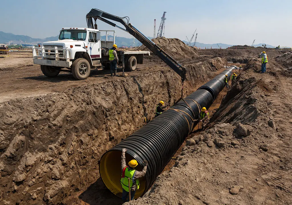 İnşaat sahasında büyük çaplı siyah HDPE koruge boru montajı: derin hendekte işçiler vinç yardımıyla büyük çaplı boru bağlantı eki yapıyor; arka planda kamyon ve dağlık arazi