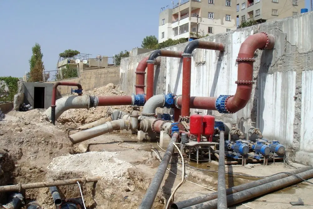 Kentsel boru bağlantı istasyonu — kırmızı çelik borular, mavi vanalar, filtreler ve pompa grubu düzeneği