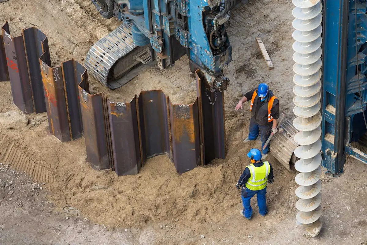 Çelik profil kazık (sheet pile) çakma — vidalı auger ve çakılmış yatay çelik profiller, iki mühendis denetim, sondaj makinesi arka planda