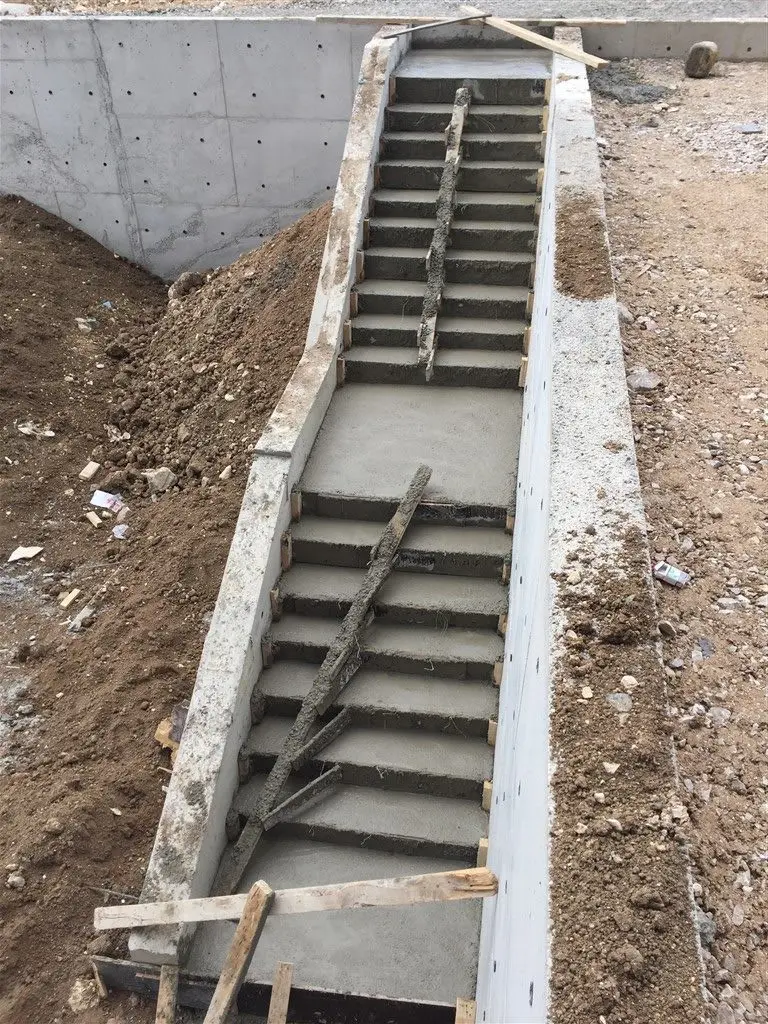 İnşaat sahasında taze beton dökülmüş tek kollu dış merdiven — ahşap kalıp kenar bordürleri görünmekte, basamaklar taze beton yüzey ile şekillendirilmiş, arka planda betonarme perde duvar mevcut