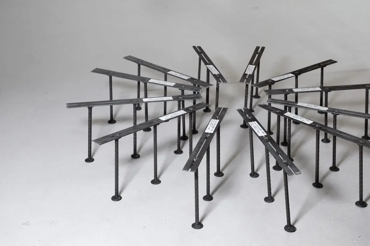 Prefabrik kayma kaması ray sistemi (shear stud rail) fiziksel örneği: çelik çubuklar ve çift başlı saplamalar radyal düzenle yerleştirilmiş, kolon çevresine monte edilmeye hazır — arka planda ikinci bir ray seti görünüyor