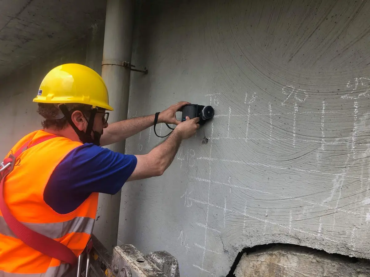 Sarı kasklı ve turuncu yelekli bir inşaat denetçisi, tamamlanmış bir betonarme yapının beton perde yüzeyine cover meter (beton örtü ölçüm cihazı) ile donatı örtü kalınlığını ölçmektedir
