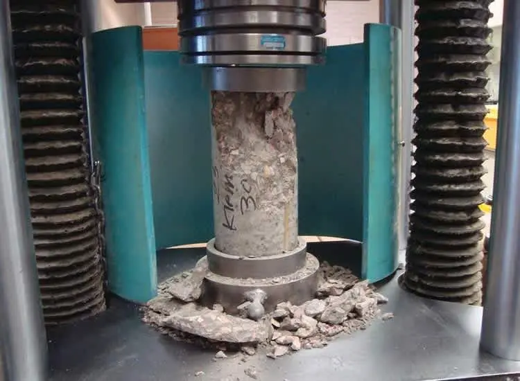 Beton basınç dayanımı deneyinde silindir beton numune, kırılma anında basınç presi arasında ezilmekte ve kırık beton parçaları tabanda birikmiş durumda görülmektedir