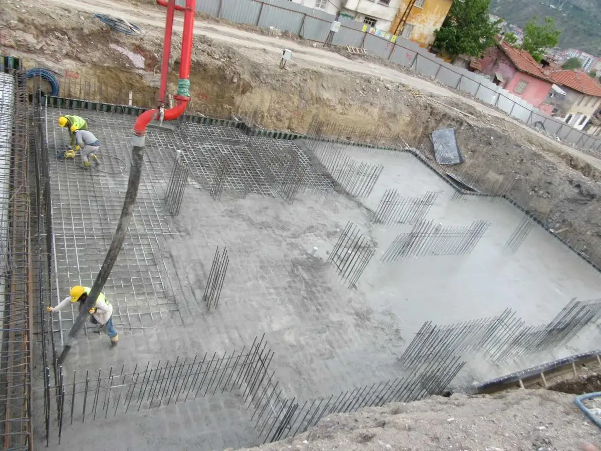Şantiyede kirişsiz döşeme beton dökümü süreci: eğimli arazide havuz yapısı temelinde açıkta donatı ve beton finişörüyle döşeme dökümü
