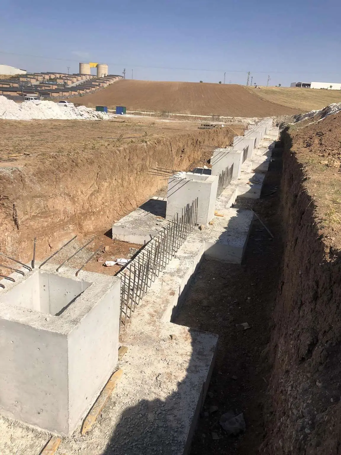 Sanayi yapısı inşaat alanında sıra halinde dizilmiş soket tipi prefabrik betonarme temeller: beton temeller yerinde döküm soket boşluklarıyla hazır, kolon montajı öncesi çelik donatılar görünüyor