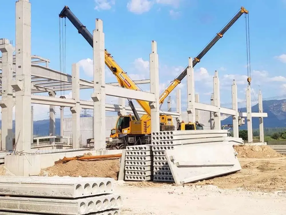 Prefabrik sanayi yapısı montajı: iki sarı vinç aynı anda boşluklu döşeme plakalarını ve kirişleri yerleştiriyor, arka planda dağlar