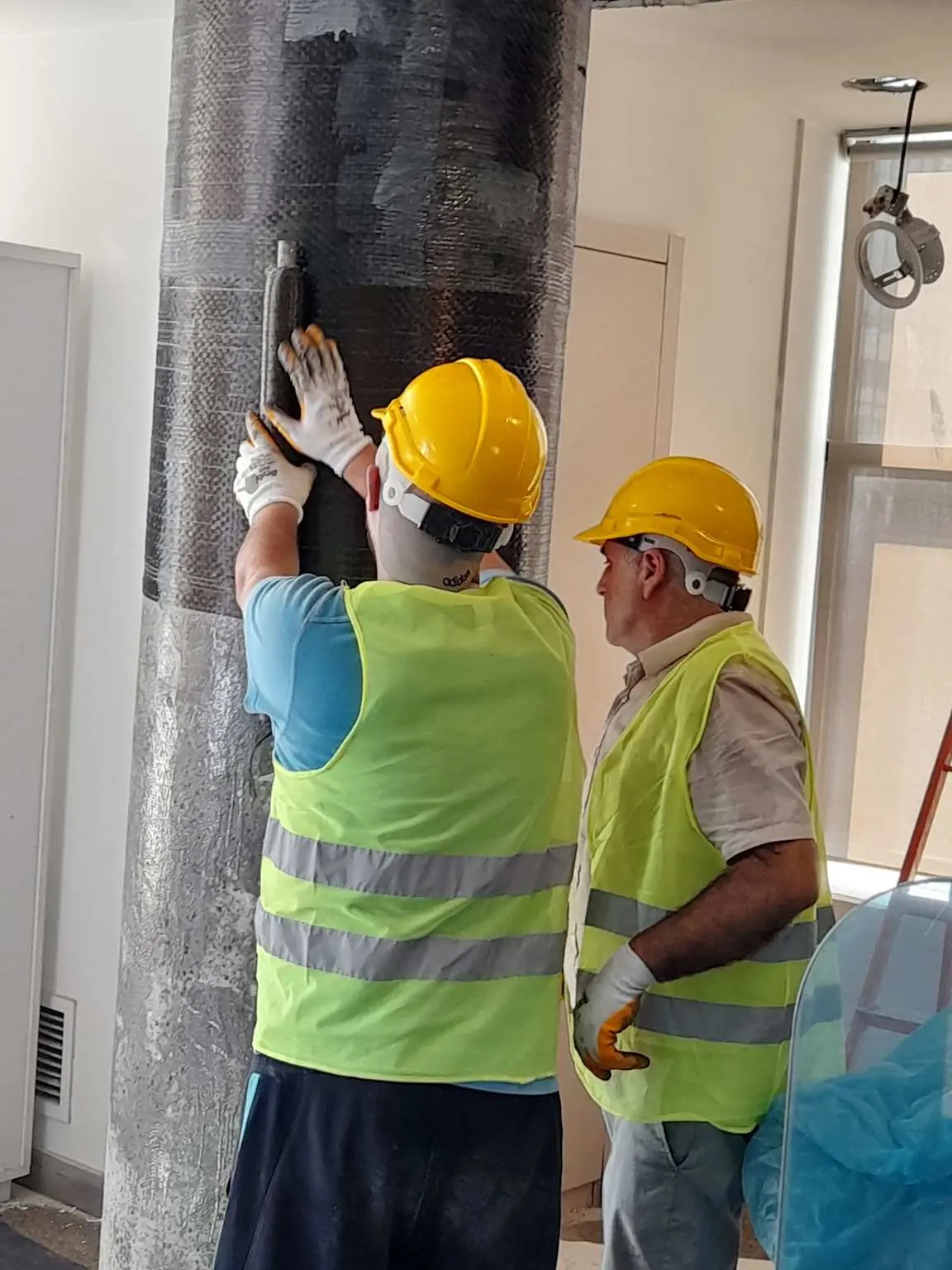CFRP karbon fiber sargı uygulaması: iki işçi bina içindeki betonarme kolona siyah karbon fiber kumaşı epoksiyle yapıştırıyor