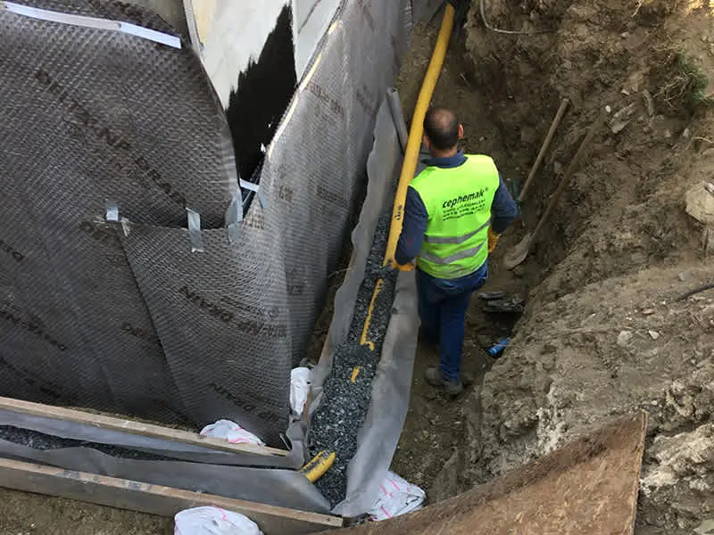 İşçi havuz cidarına su yalıtım membranı uygularken: siyah membran plakaları ile kapı açıklığı kalıbı arasında drenaj çakıl serimi, inşaat güvenlik yeleği
