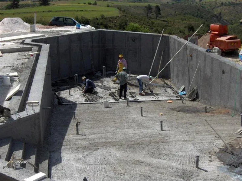 Büyük betonarme havuz inşaatında taban beton dökümü: işçiler ve ekipmanla beton serimi, kalıp sistemi ve başlangıç çubuklarından oluşan geniş havuz yapısı
