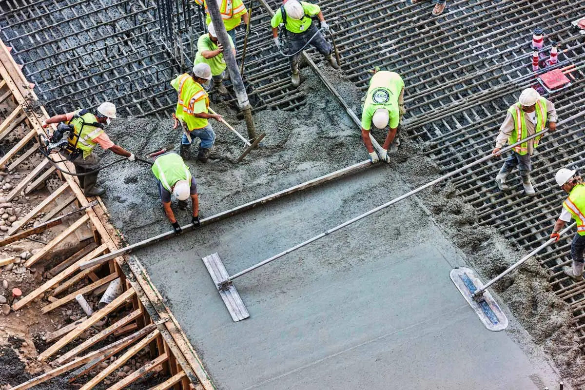 Açık havada inşaat sahasında taze dökülen betonarme döşeme — sarı yelek ve baret giyen altı kişilik ekip, eğim rayı üzerinde beton perdahı ve vibrasyonu gerçekleştiriyor