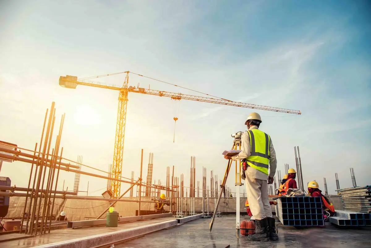 İnşaat şantiyesinde kule vinci ve beton kalıplı kolonlar arasında theodolite ile ölçüm yapan sarı yelek ve baret giyen harita mühendisi