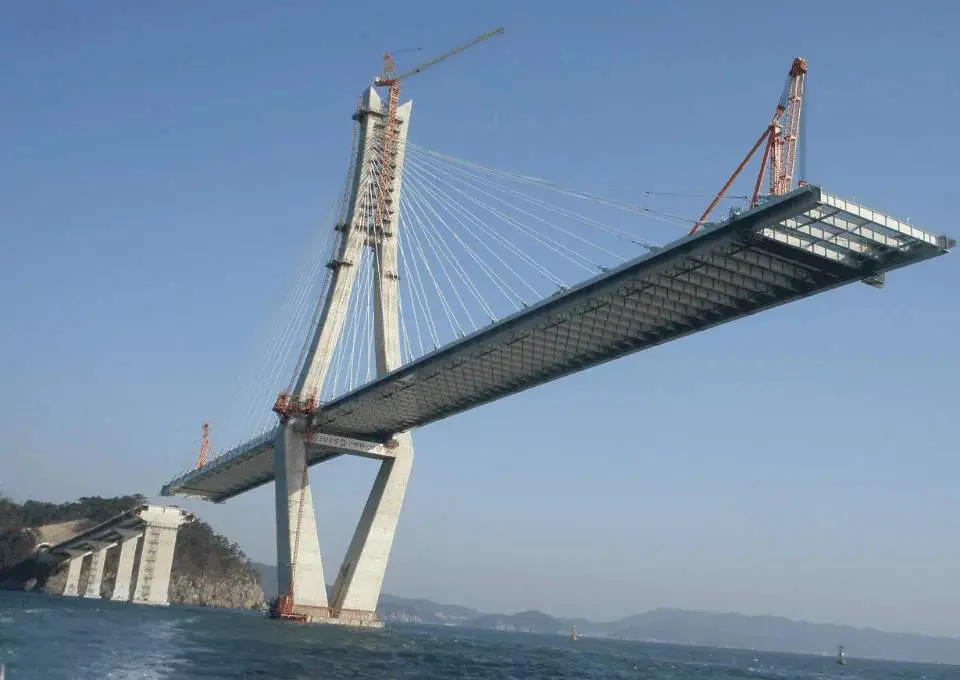 Eğik kablo köprüsü inşaatı denizden görünüm — iki beton pylon, kısmi tamamlanmış tabliye stay kablolarıyla asılı