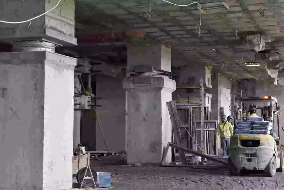 İnşaat halindeki yapının yalıtım katı: beton kolonlar üzerinde yerleştirilen sismik izolatör (ortada) ile çevresindeki kalıp ve altyapı çalışmaları; işçiler ve forklift ile depo/inşaat ortamı.