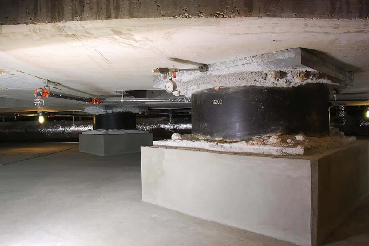 Yapının bodrum katında sismik izolasyon katmanı: beton kolonlar üzerine yerleştirilmiş büyük kauçuk izolatörler (siyah silindirik elemanlar, 11200 etiketli), bağlantı boruları ve tavan sistemi görülmektedir.