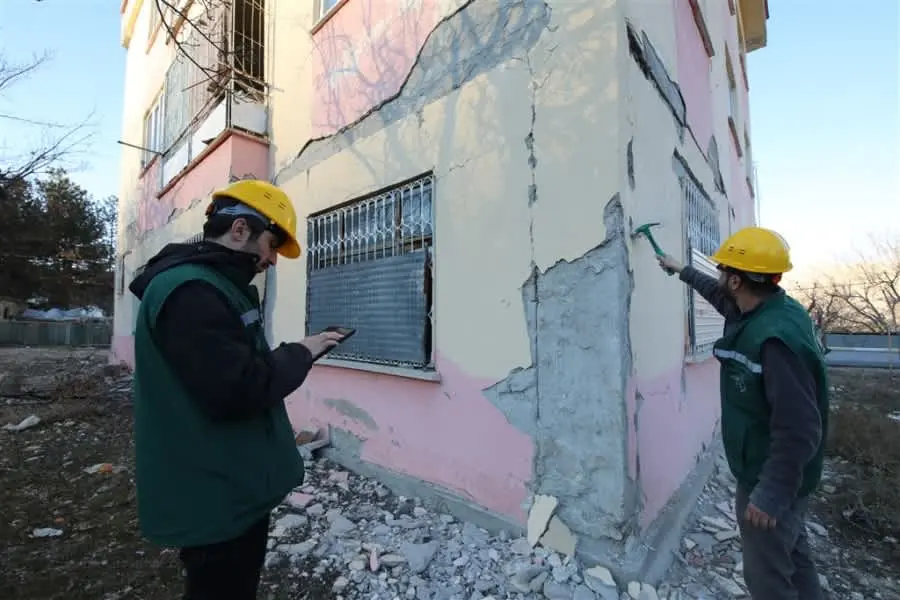 İki mühendis baret takarak çatlak dökülmüş bina köşesinde hasar tespiti yapıyor: biri tablet ile form dolduruyor, diğeri çekiç ile beton kontrol ediyor