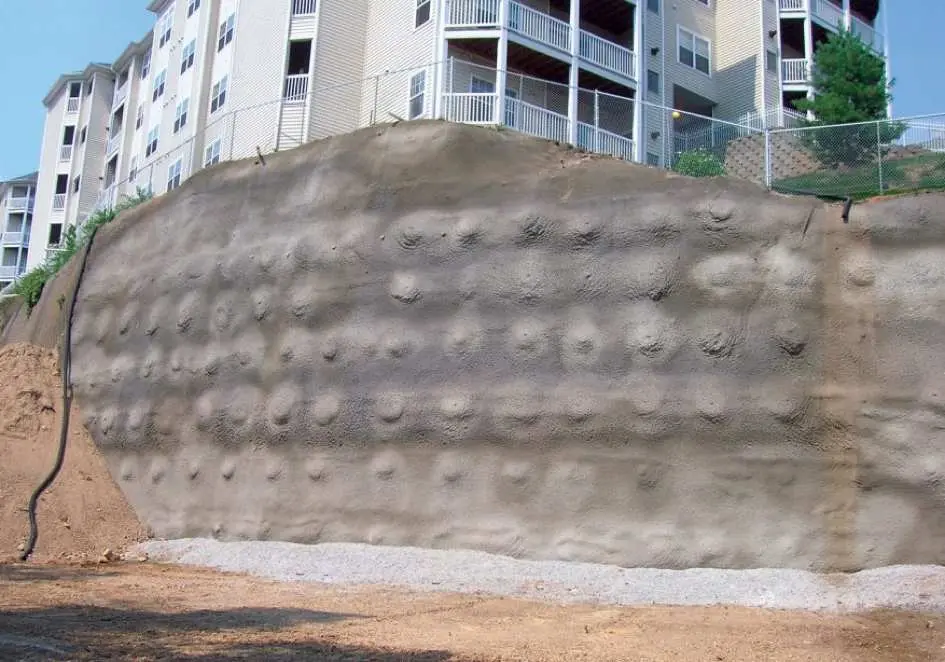 Zemin çivisi uygulanmış shotcrete yüzey duvarı: beton yüzeyde düzenli aralıklarla nail başları görülmekte, arkada apartman binası