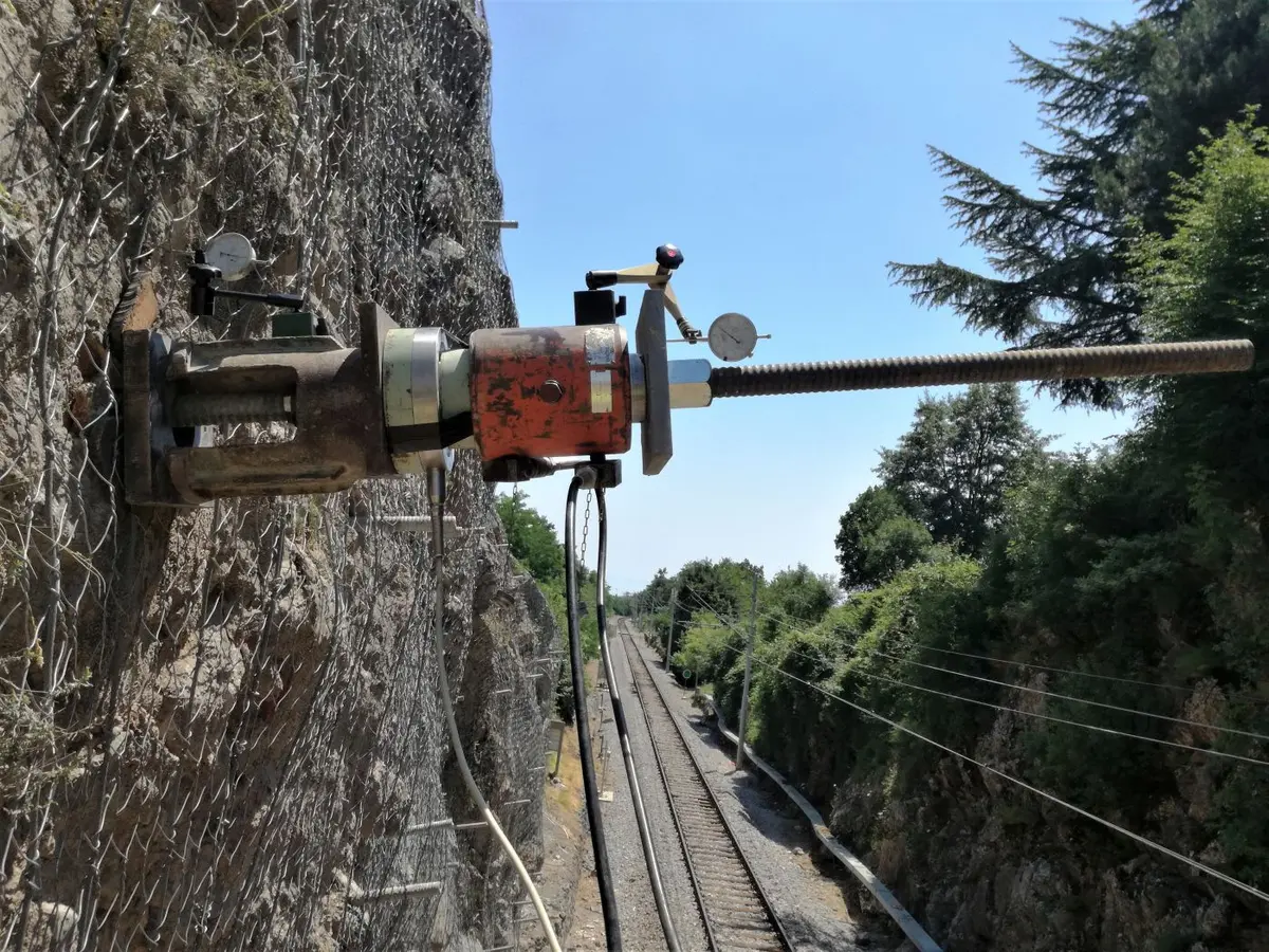 Kayalık şev yüzeyine monte edilmiş kırmızı renkli ankraj delgi ve germe ekipmanı — demiryolu hattı yanındaki kaya şev sabitleme çalışması