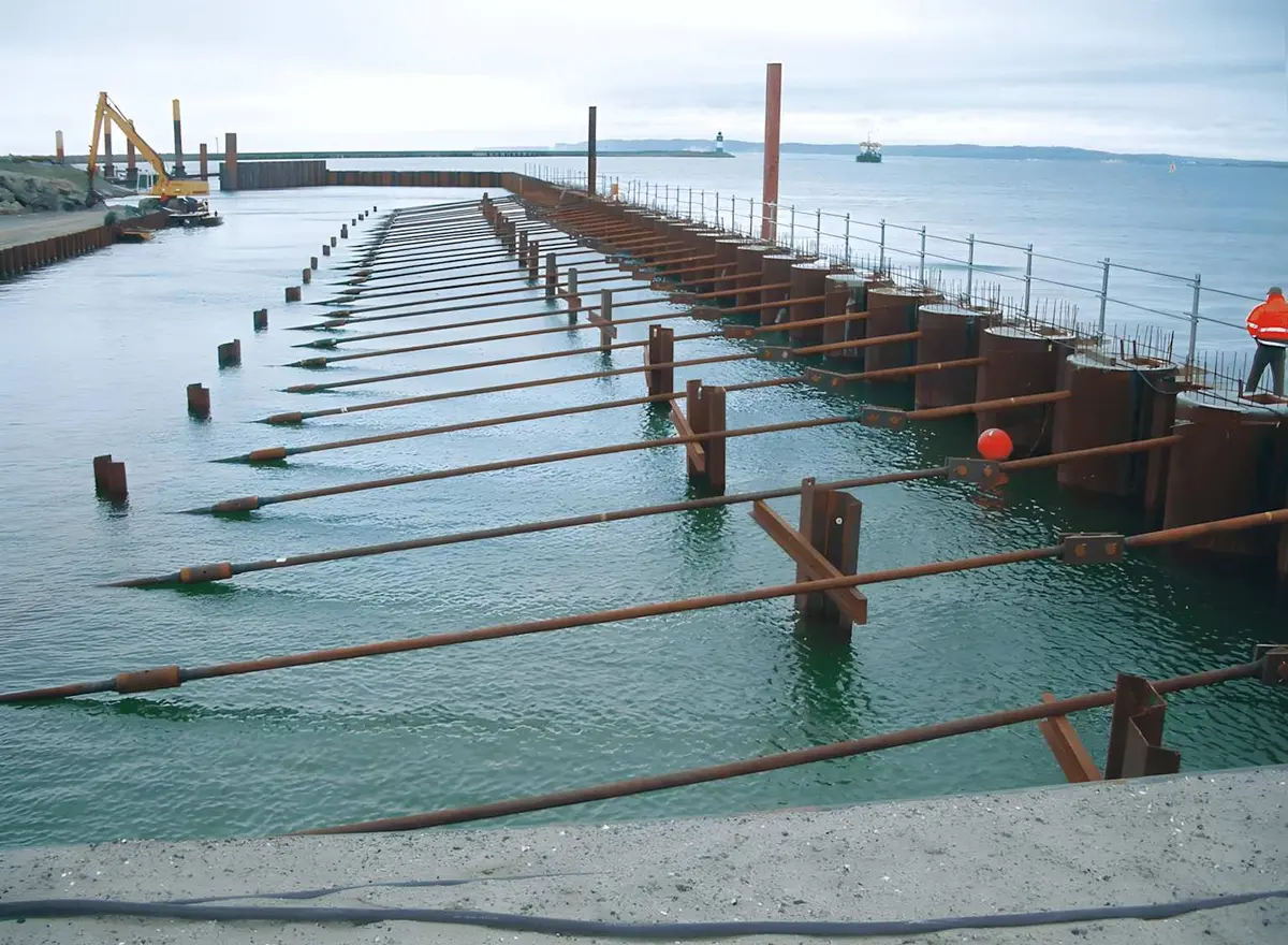 Rıhtım yapısında ankrajlı palplanş sisteminin havadan görünümü — çelik palplanş profiller su içinde dikey dizili, çok sayıda yatay tie rod (ankraj çubuğu) deniz tabanına uzanıyor, sarı vinç uzakta görünüyor