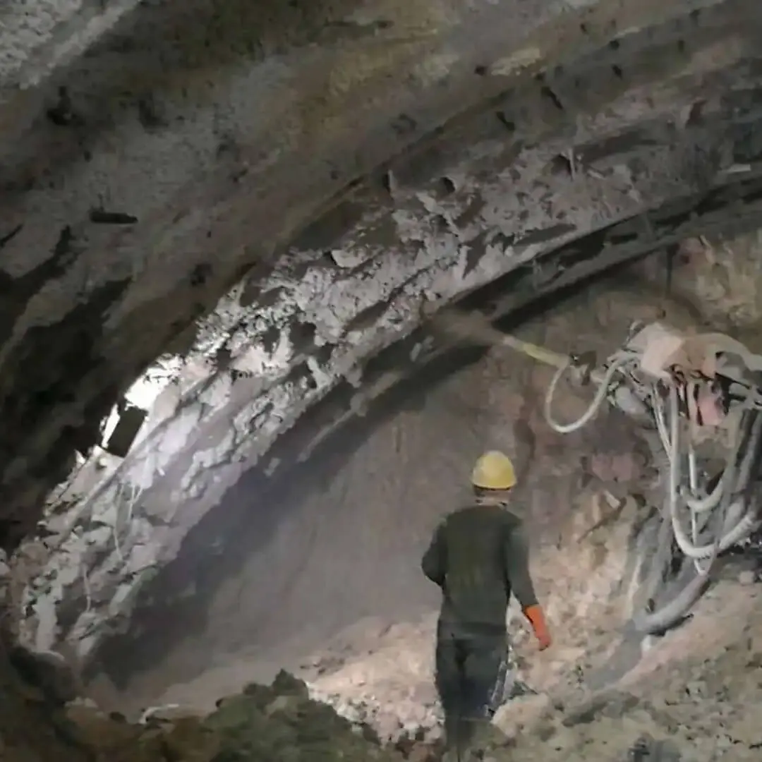 Türkiye kaya tünelinde NATM yöntemiyle shotcrete uygulaması — tünel içinde işçi görünüyor, tavan ve yanlarda gri beton püskürtme izleri var, kaya bulonu hortumları asılı, yoğun kayaçlı tavan