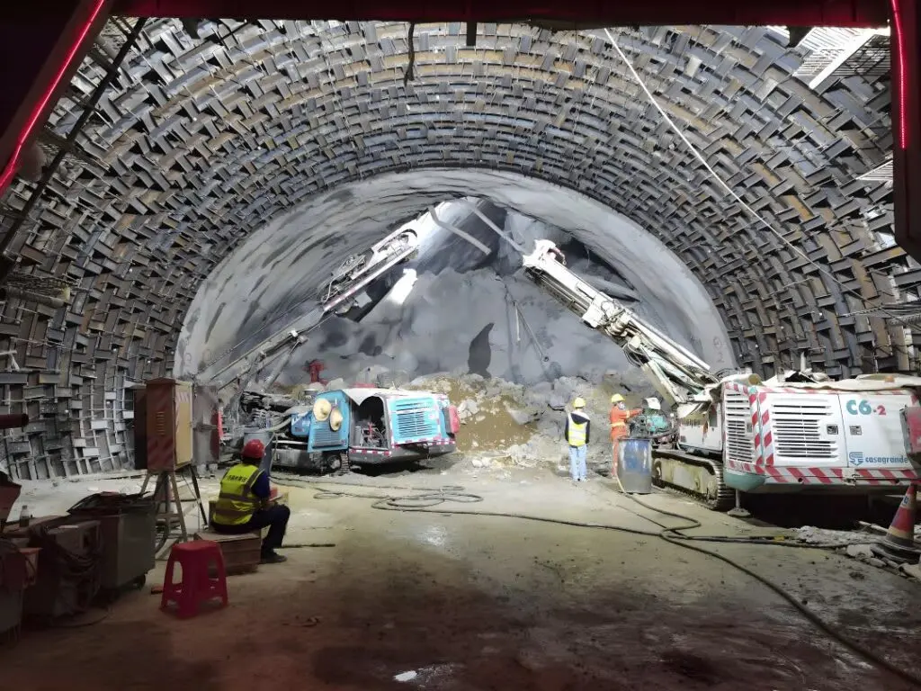 Büyük çaplı NATM tünel kazısı — çift kollu jumbo delgi makinesi, shotcrete uygulanmış tünel yüzeyi, çelik kemer görünümü