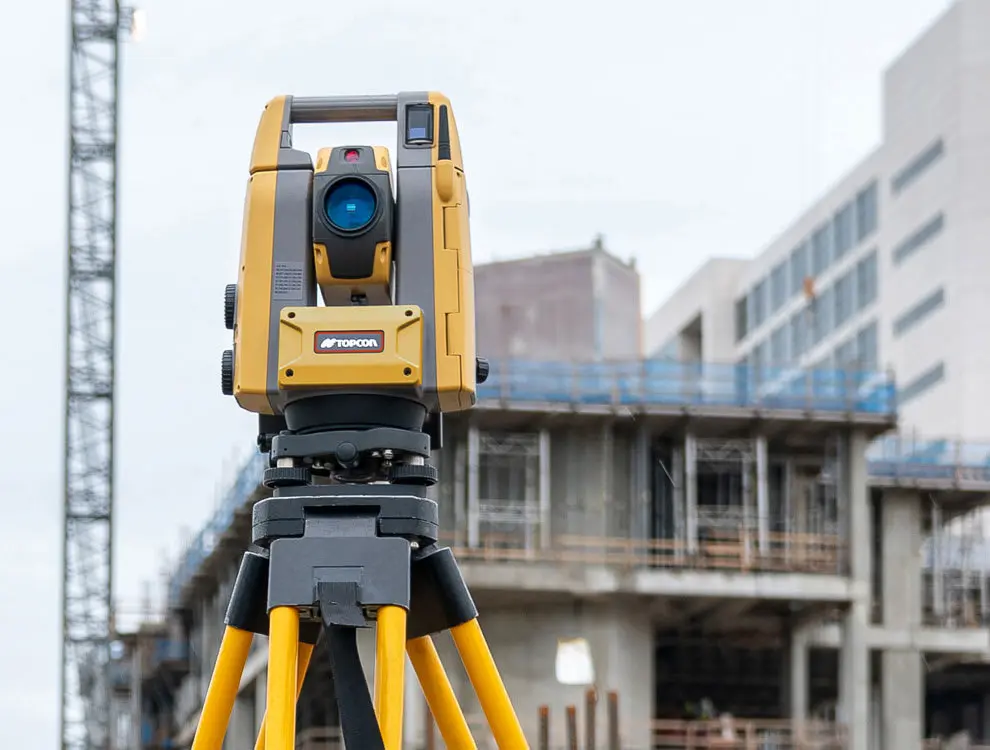 Topcon total station şantiye ortamında inşaat binası önünde tripod üzerinde, sarı gövde ve mavi EDM mercek görünmekte