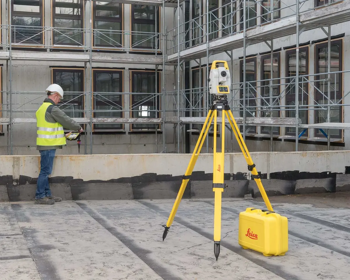 Leica total station şantiyede tripod üzerinde kurulu, arka planda sarı yelekli teknisyen ölçüm yapıyor, inşaat iskelet yapısı görünmekte