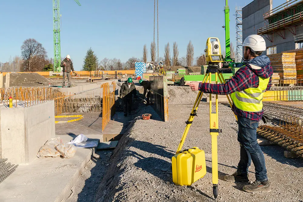 Şantiyede temel hafriyatı kenarında Leica total station kurulu, mühendis beton zemin üzerinde çalışıyor, arka planda inşaat vinci ve işçiler görünmekte
