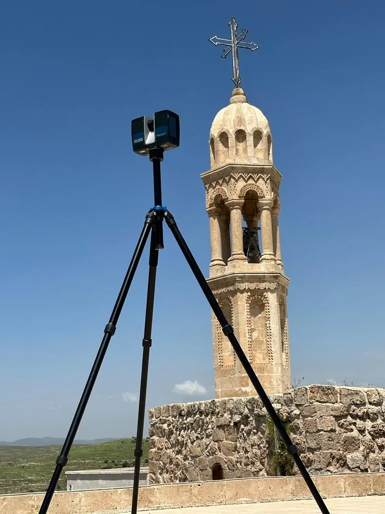 Tarihi kilise kulesi önünde tripod üzerinde kompakt lazer tarayıcı; mavi gökyüzü ve taş kule görünüyor