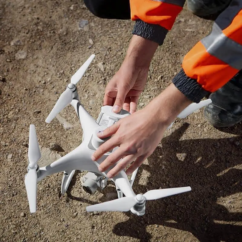 Turuncu reflektif yelek giyen mühendis İHA'yı inşaat sahasında test ediyor; beyaz multirotor İHA çakıllı zeminde hazırlanıyor