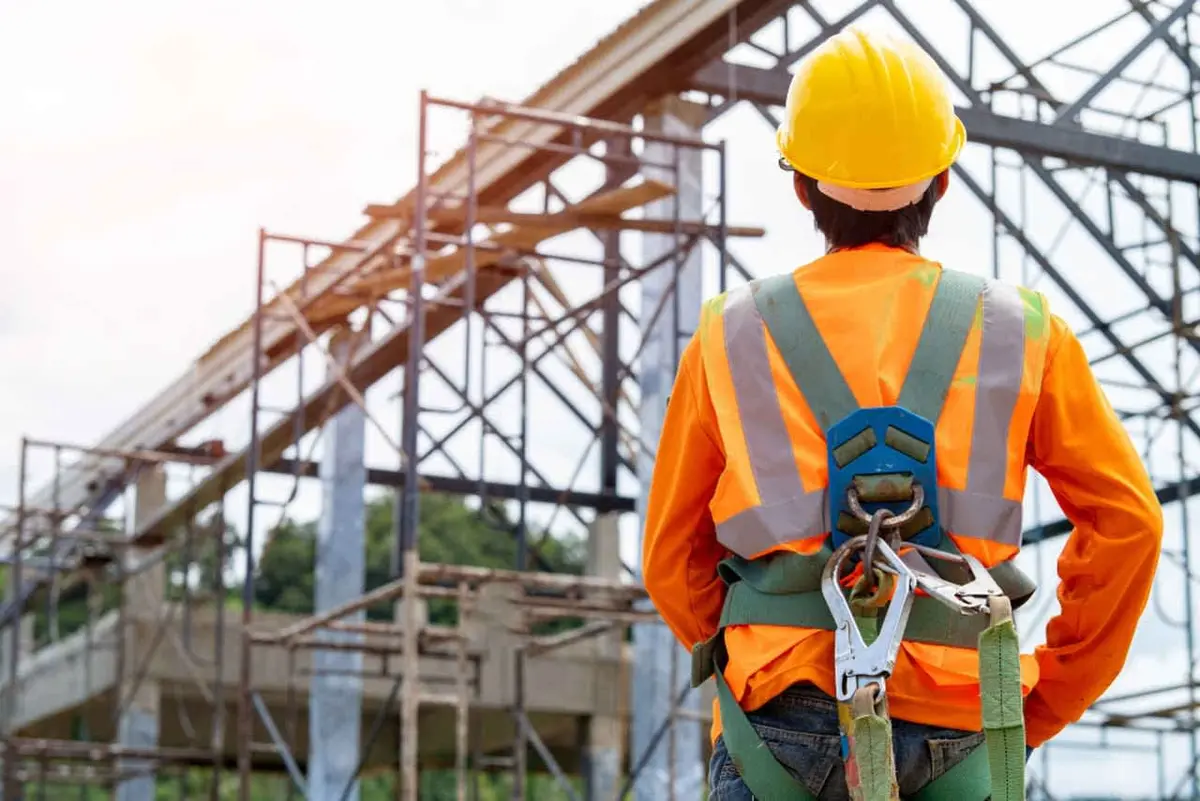 İnşaat şantiyesinde emniyet kemeri giyen işçi, arka planda çatı makası ve iskele yapımı — yüksek risk ortamında KKD kullanımı