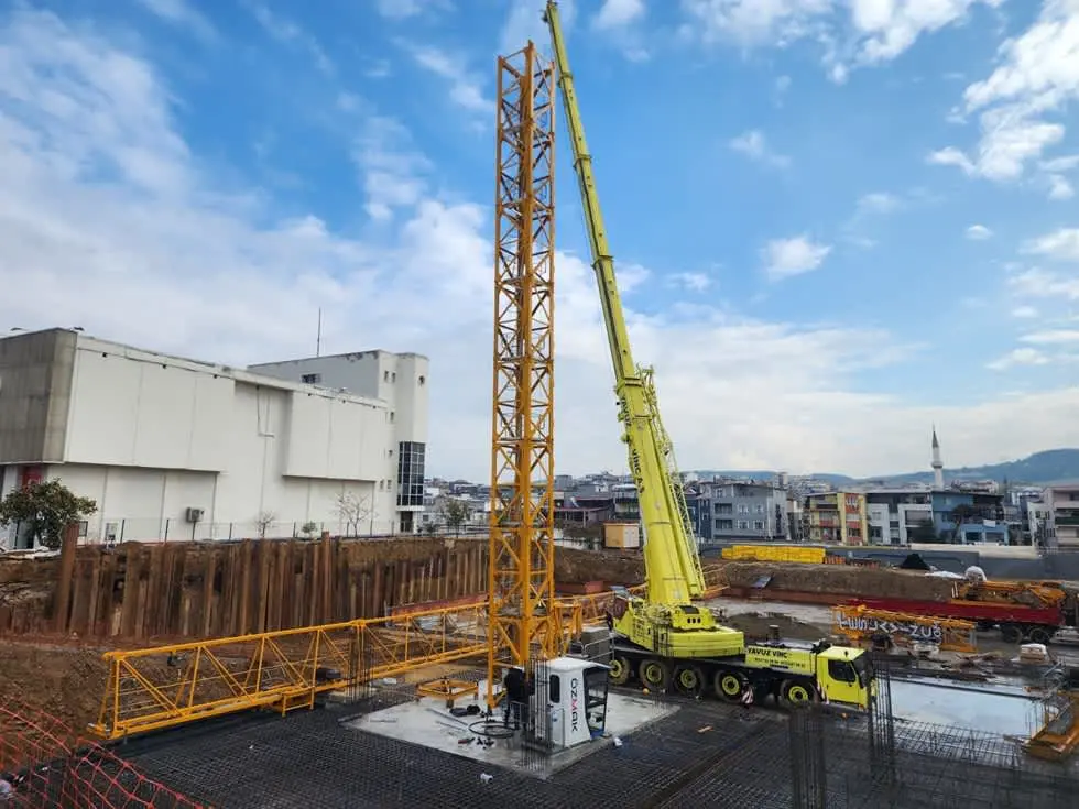 İnşaat şantiyesinde kule vinci yükseltirken yardımcı olarak kullanılan sarı renkli teleskopik mobil vinç; arka planda apartman binaları