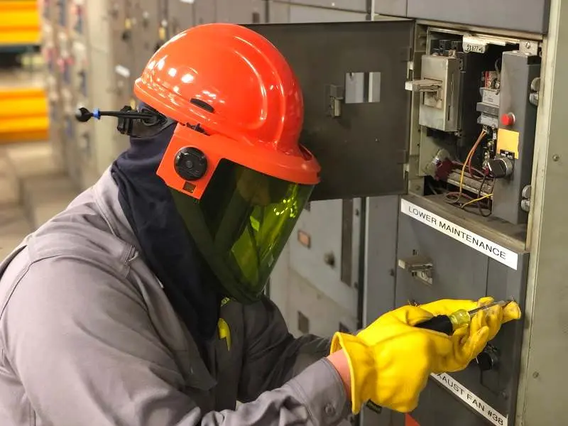 Elektrik bakım personeli arc flash KKD kullanıyor — turuncu baret ve açık yeşil vizörlü yüz siperi, ark korumalı gri elbise ve sarı yalıtkan eldiven; açık elektrik panosu önünde tornavida ile çalışma