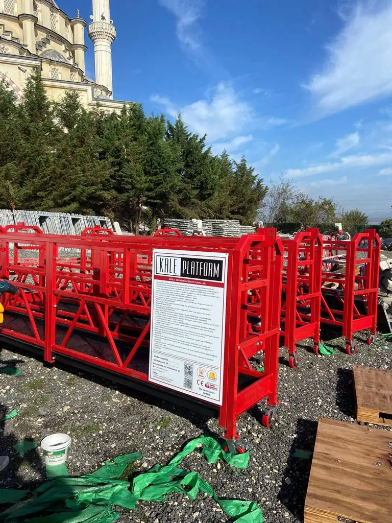 CE belgeli kırmızı renkli asılı iskele (suspended scaffold) platformları: cami şantiyesi önünde depolanmış, her iki yanında yükseltici motorları bulunan alüminyum platformlar