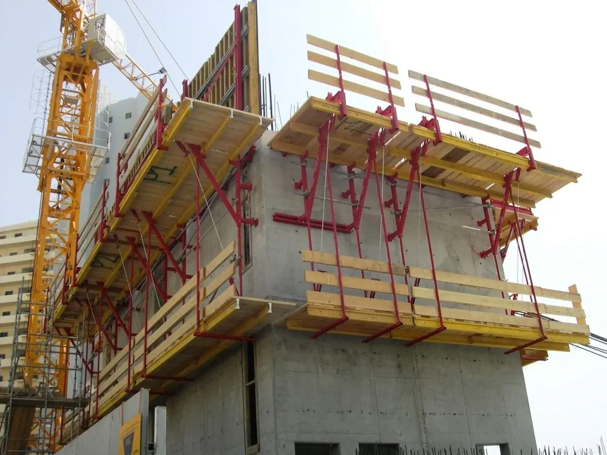 Bina köşesinde kurulu kırmızı çelik tırmanır kalıp konsolları ve ahşap platformlar; arka planda sarı inşaat vinci görülmekte